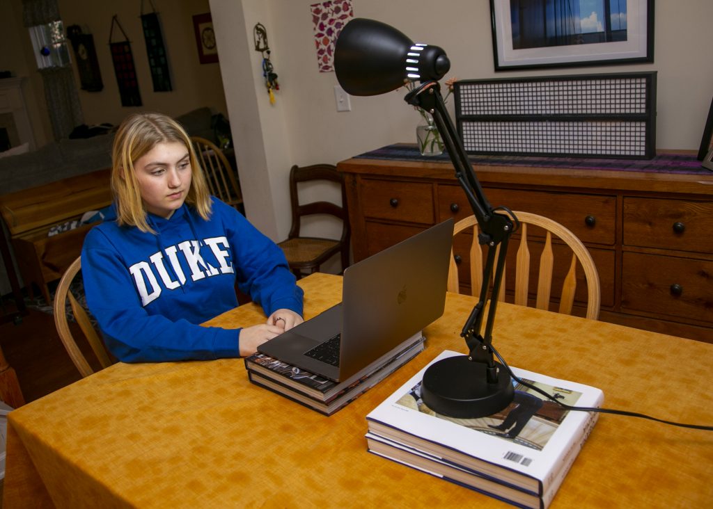 The image depicts a young woman sitting at a kitchen table in front of a laptop.